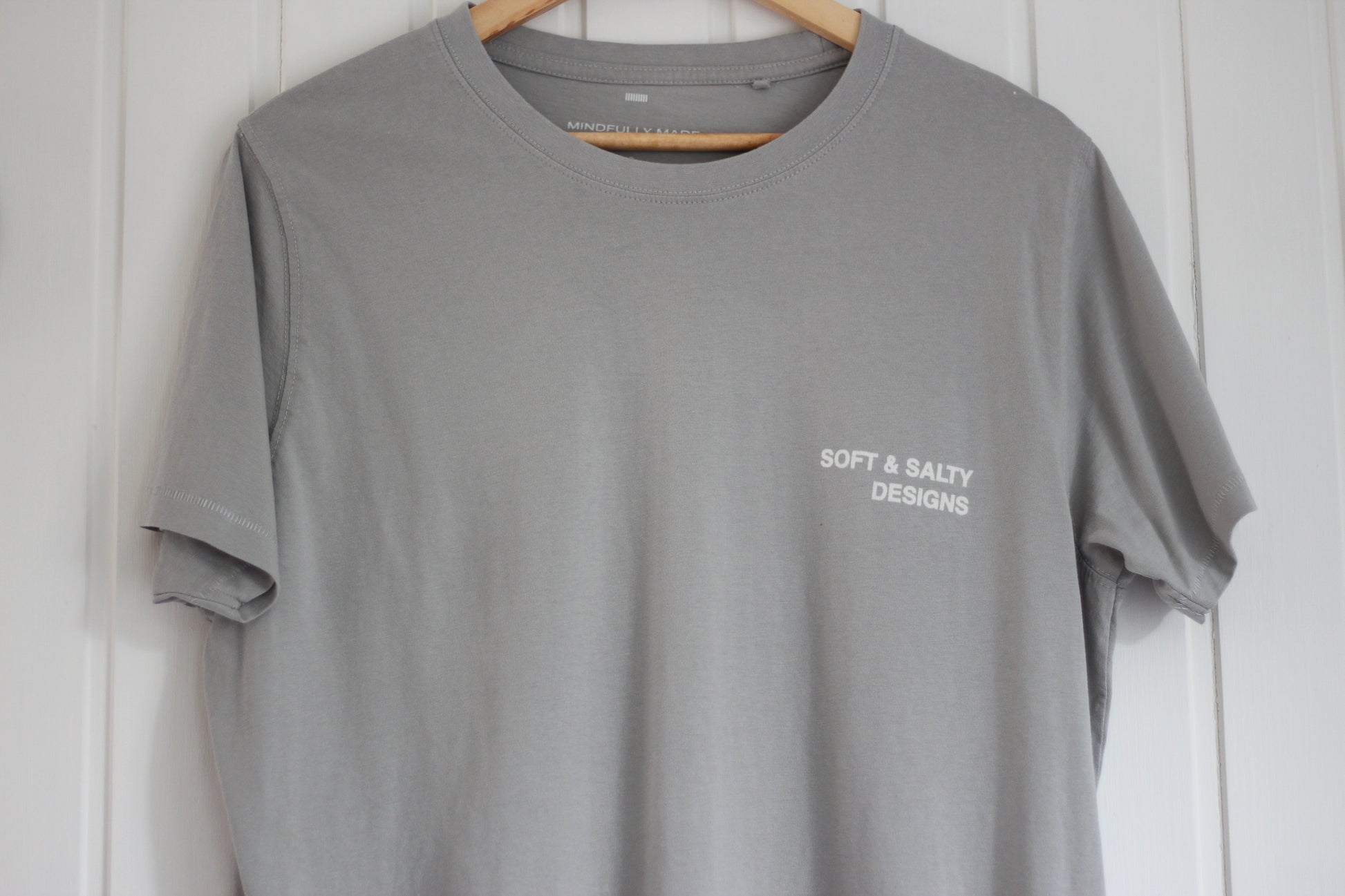 Gray t-shirt with 'Soft & Salty Designs' logo on a wooden hanger against a white wall.
