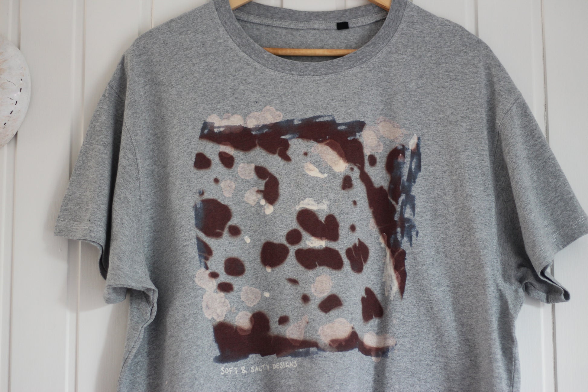 Grey t-shirt with kelp printed design on a wooden hanger against a white wall.