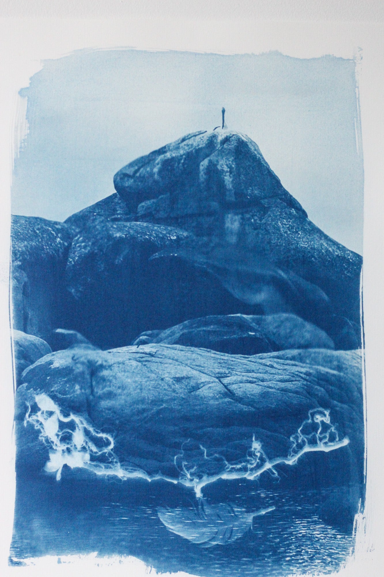 A monochrome blue-toned landscape artwork showing a rocky coastal formation with a pelican sitting on the highest rock above water, rendered in a cyanotype-style print with rough white margins.