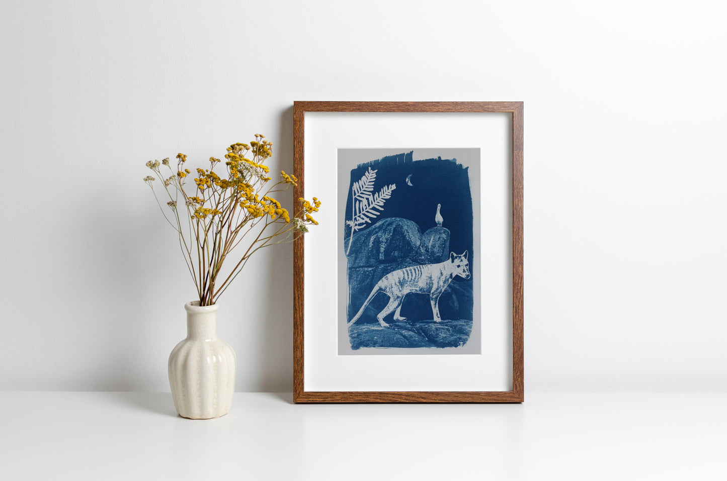 A framed art print featuring a blue and white illustration of a thylacine in a forest, styled on a white surface next to a small white vase with yellow flowers against a white wall.