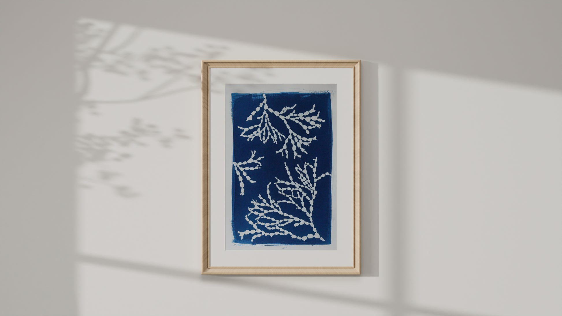 A framed art print featuring white coral or sea-plant motifs on a deep blue background, mounted on a light gray wall with soft shadow projections.