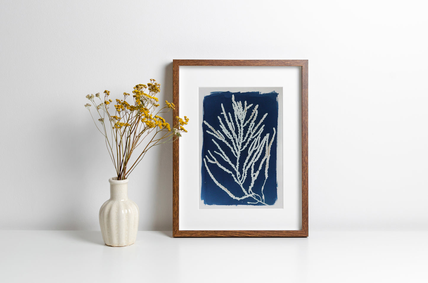 A framed blue and white coral print in a brown wooden frame, styled on a white surface next to a small white vase with yellow dried flowers against a white wall.