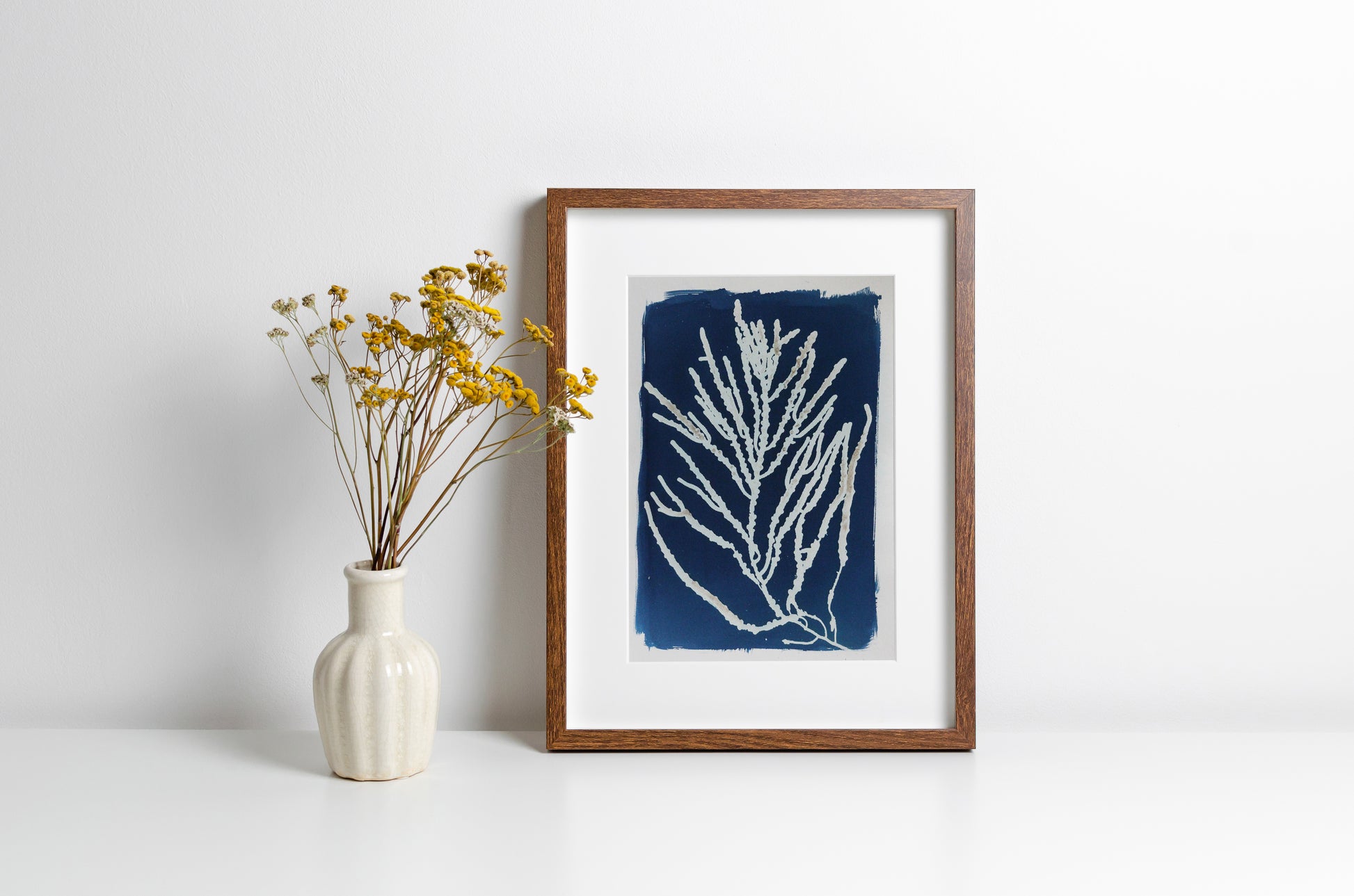 A framed blue and white coral print in a brown wooden frame, styled on a white surface next to a small white vase with yellow dried flowers against a white wall.