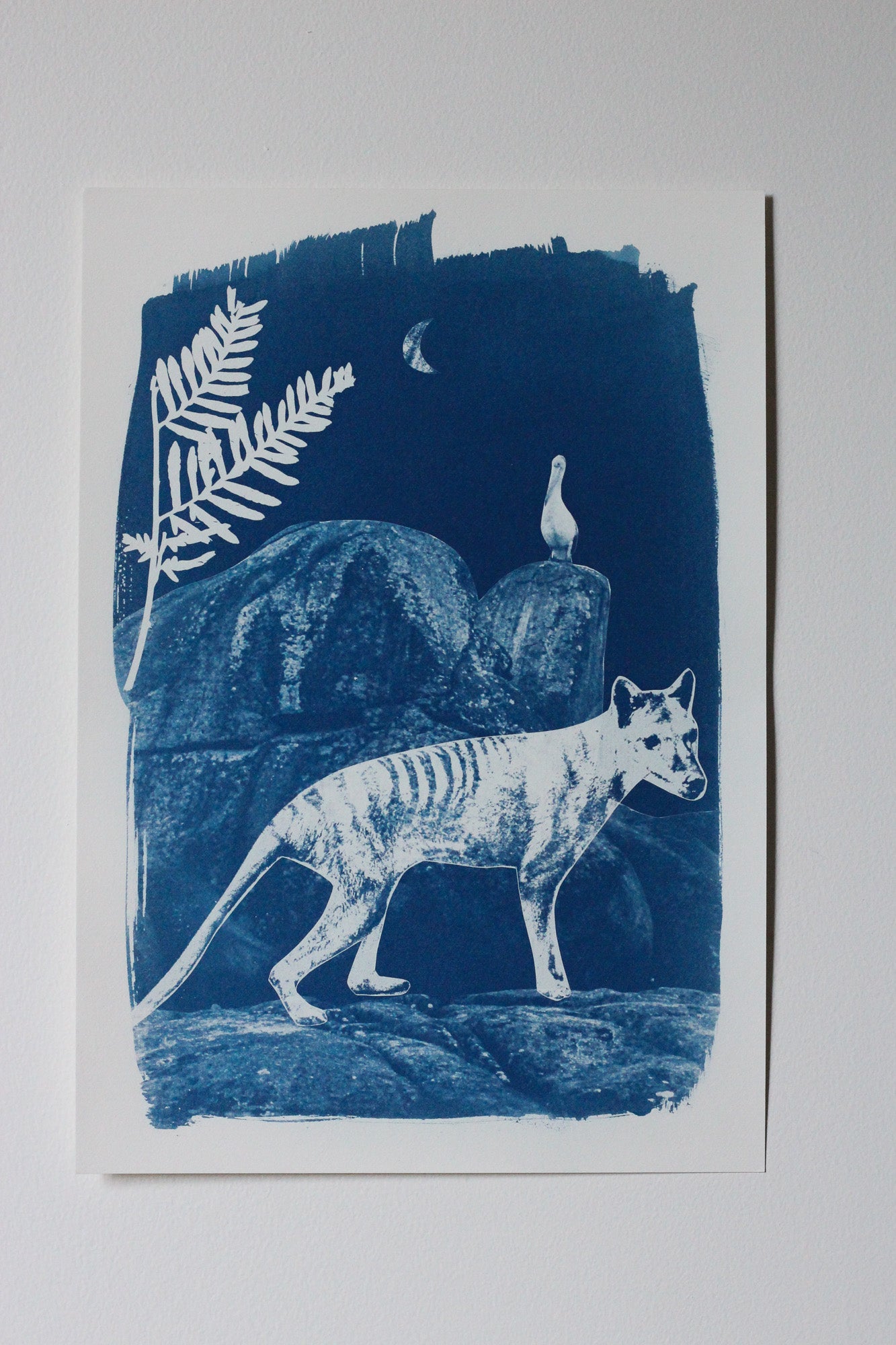 A blue and white art print depicting a Tasmanian tiger walking in a rocky landscape at night, with a fern leaf and a pelican on a rock in the background, shown against a neutral light gray background.