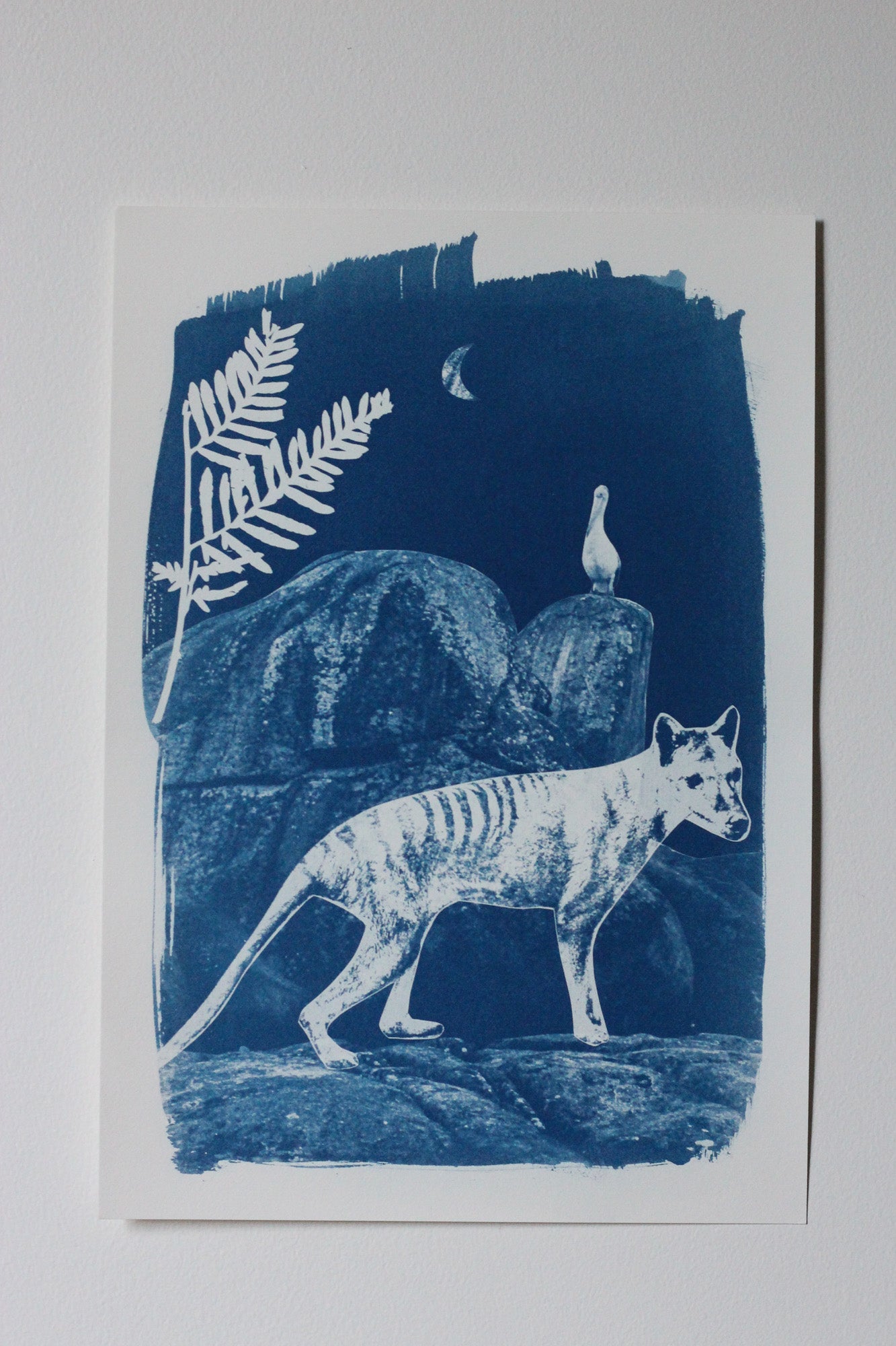 A blue and white art print depicting a Tasmanian tiger walking across rocks, with a fern leaf and a pelican on a rock in the background, photographed against a neutral light gray background.