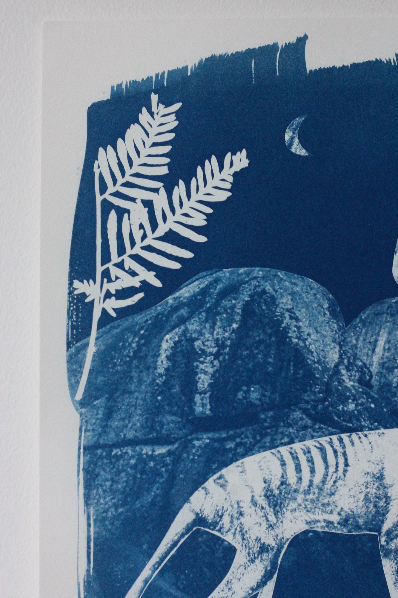 A close-up of a blue and white art print featuring a fern leaf, a thylacine, a crescent moon, and abstract rock-like forms.