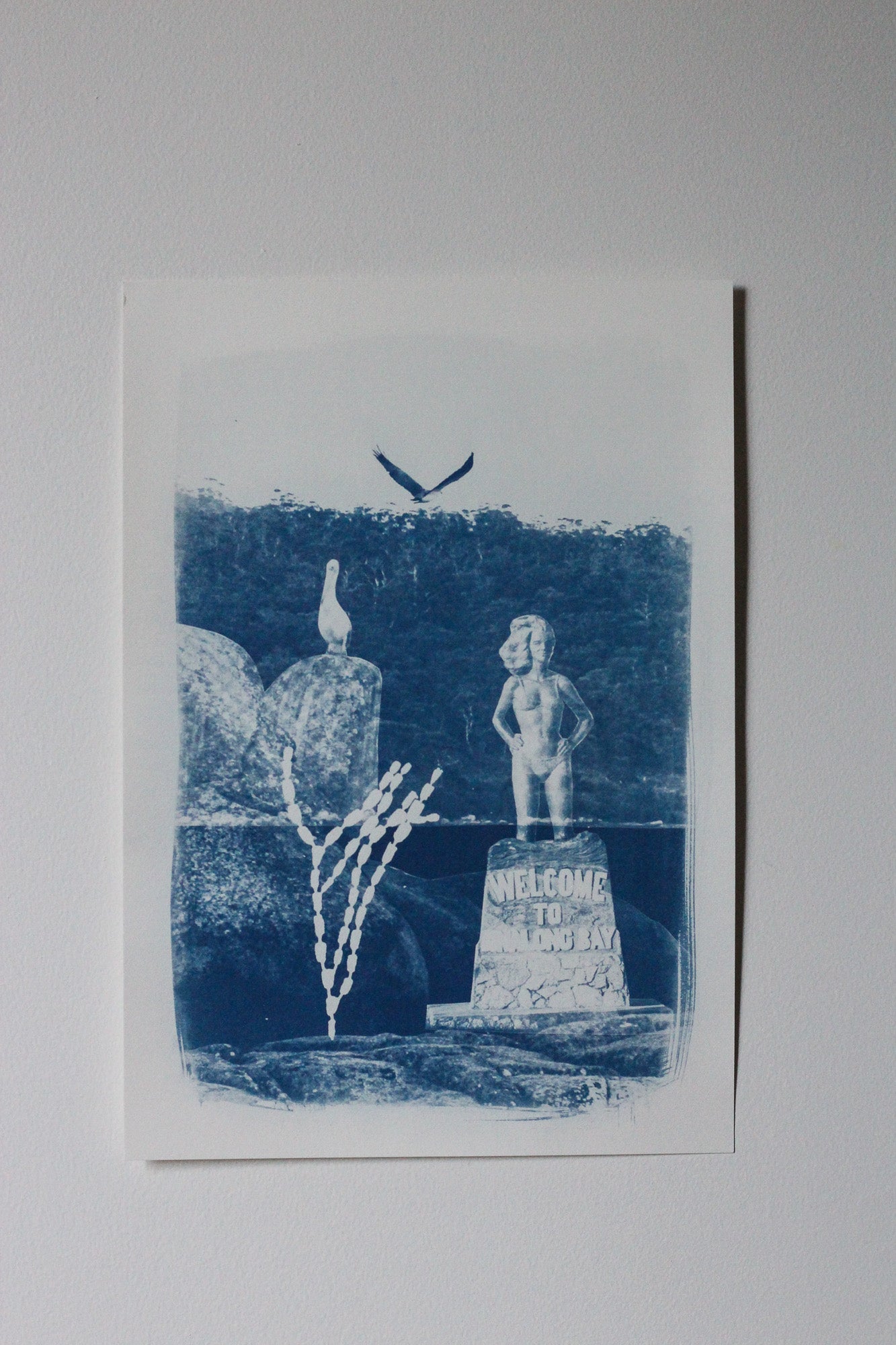 A blue and white art print or postcard featuring a stylized scene with a bird, a statue, rocks, and a sign reading 'WELCOME TO BINALONG BAY'.