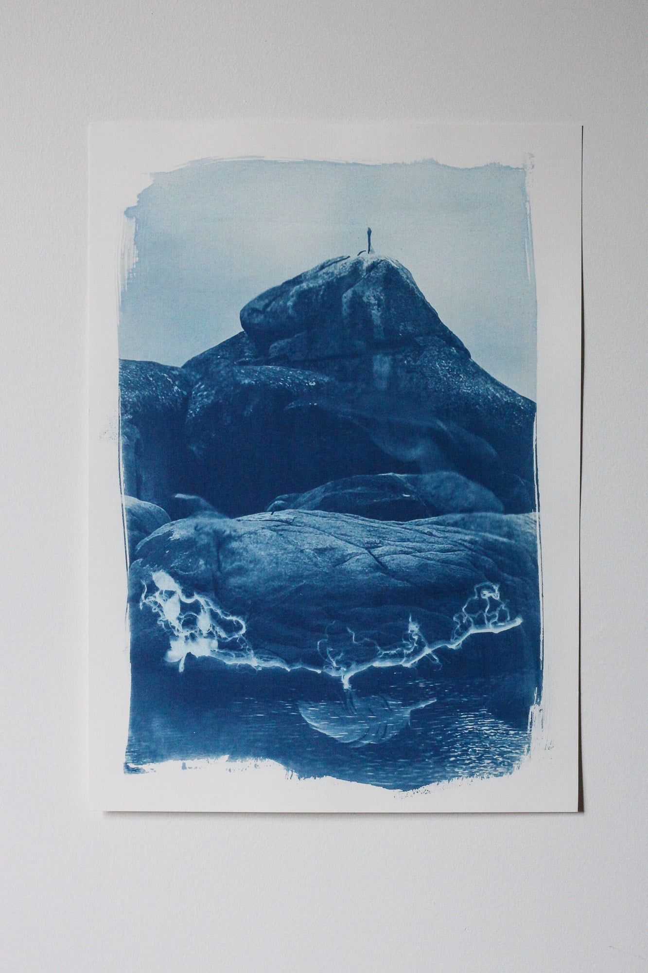 A cyanotype-style art print depicting a pelican sitting atop rocky landscape with blue tones on white paper, photographed against a light neutral background.