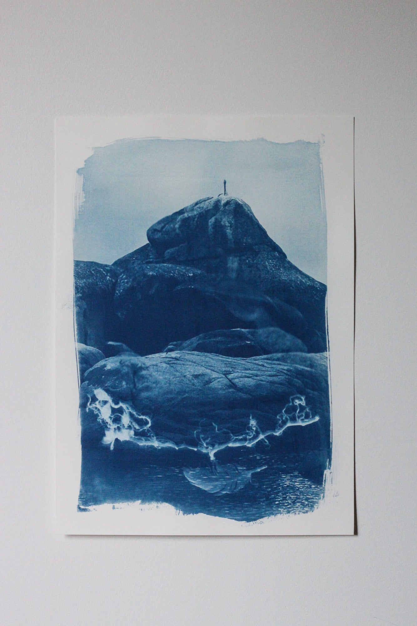 A cyanotype-style art print depicting a pelican sitting atop rocky landscape with blue tones on white paper, photographed against a light neutral background.