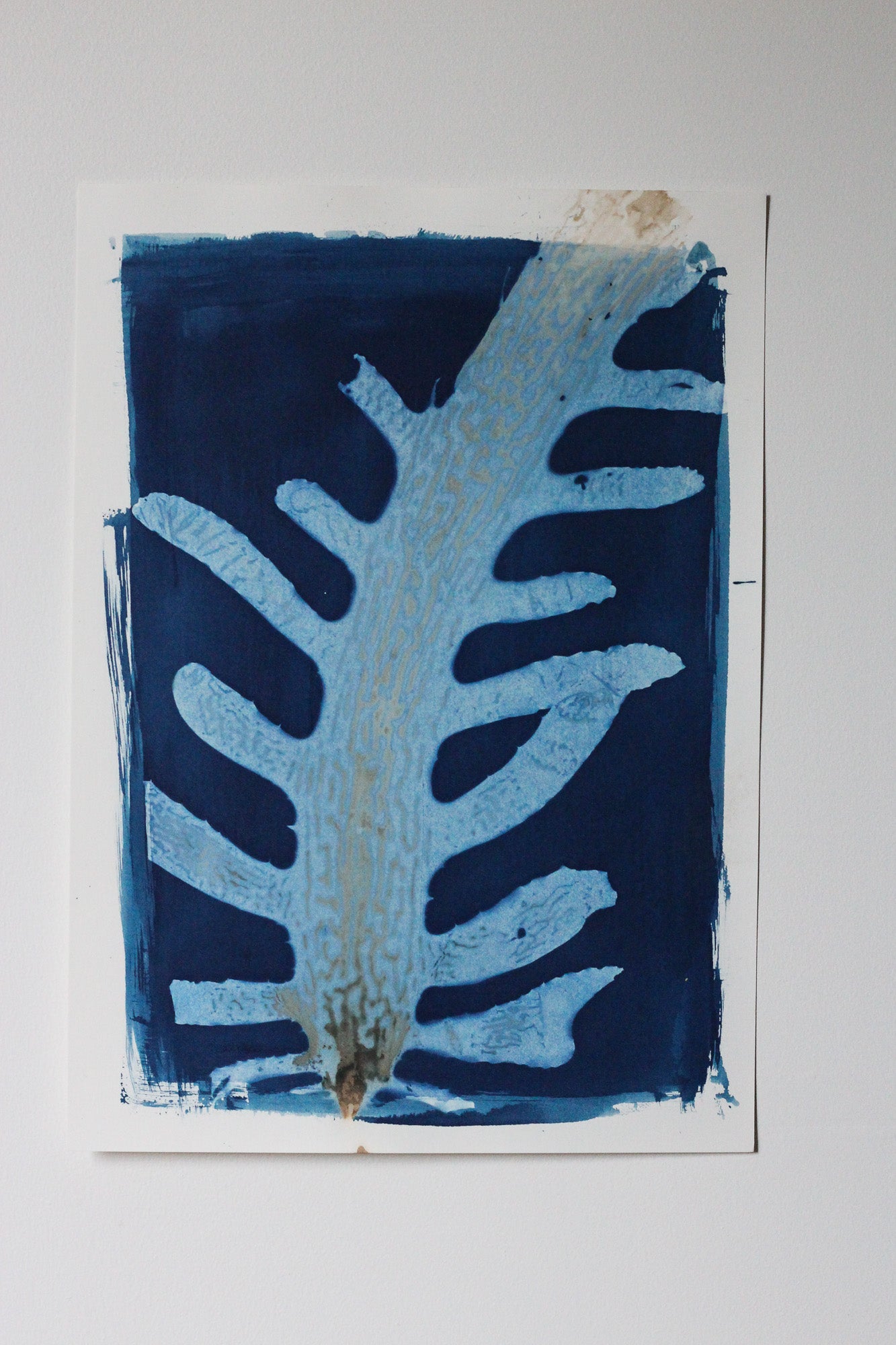 An abstract art print featuring a stylized sea weed motif in light blue and white on a dark navy blue background, mounted on a white paper sheet against a neutral light gray wall.