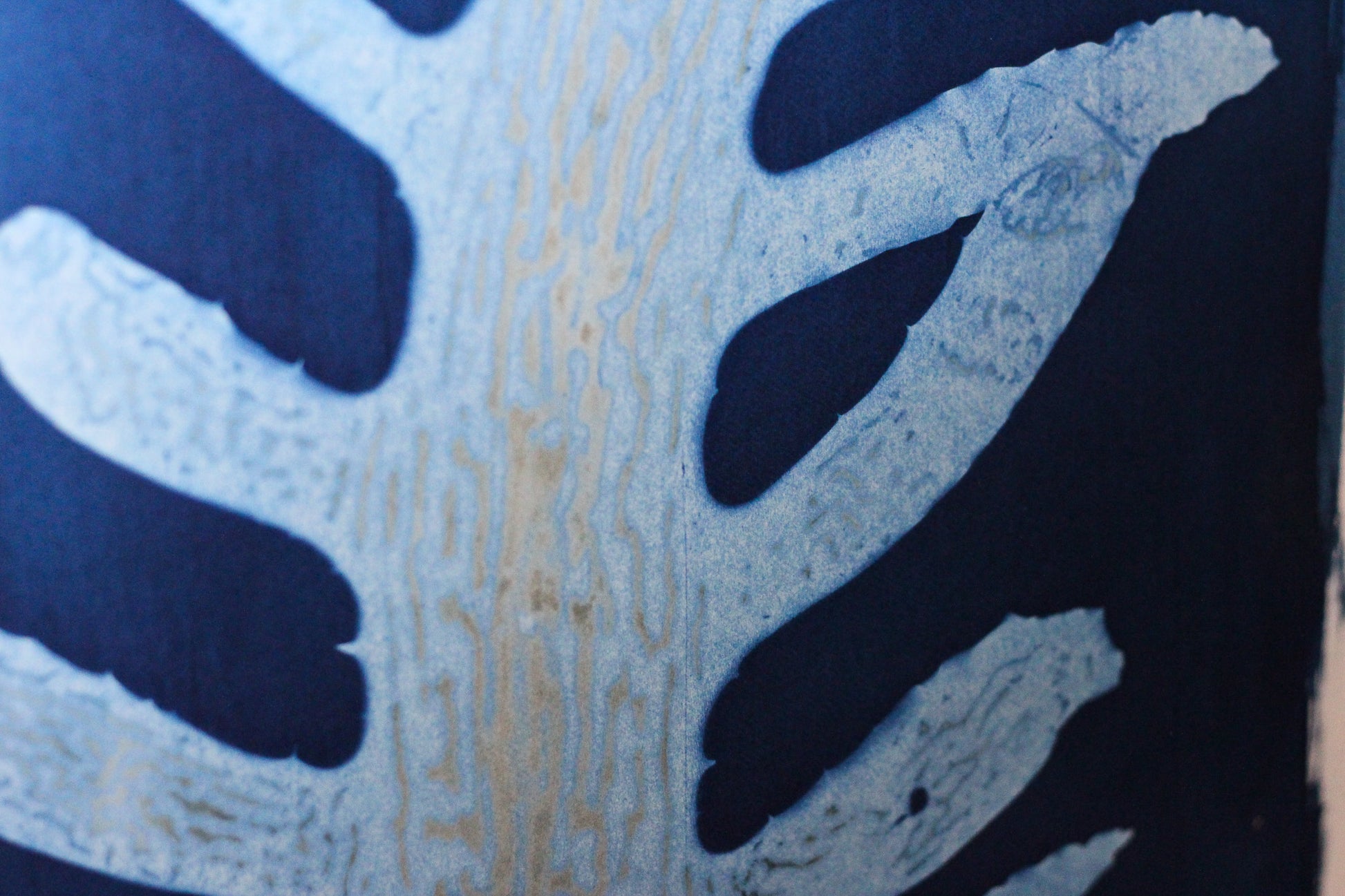 A close up of an abstract cyanotype art print featuring a seaweed organic pattern in light blue and dark navy blue on a white sheet of paper, photographed against a plain white wall.