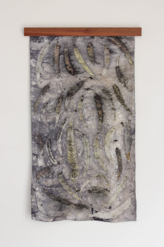 A bundle dyed wall hanging featuring eucalyptus leaf prints hanging from a wooden frame on a white background. 