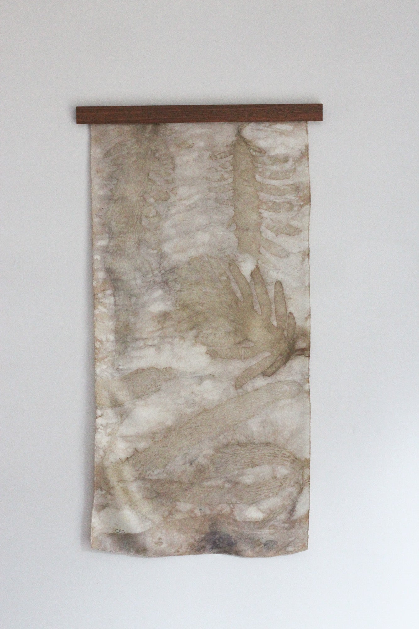 A vertical fabric wall hanging or tapestry with an abstract watercolor-style design in beige, brown, and gray tones, suspended from a dark wooden rod against a plain light gray wall.