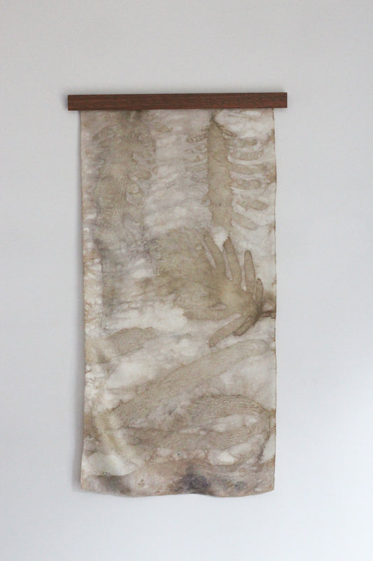 A vertical fabric wall hanging or tapestry with an abstract watercolor-style design in beige, brown, and gray tones, suspended from a dark wooden rod against a plain light gray wall.