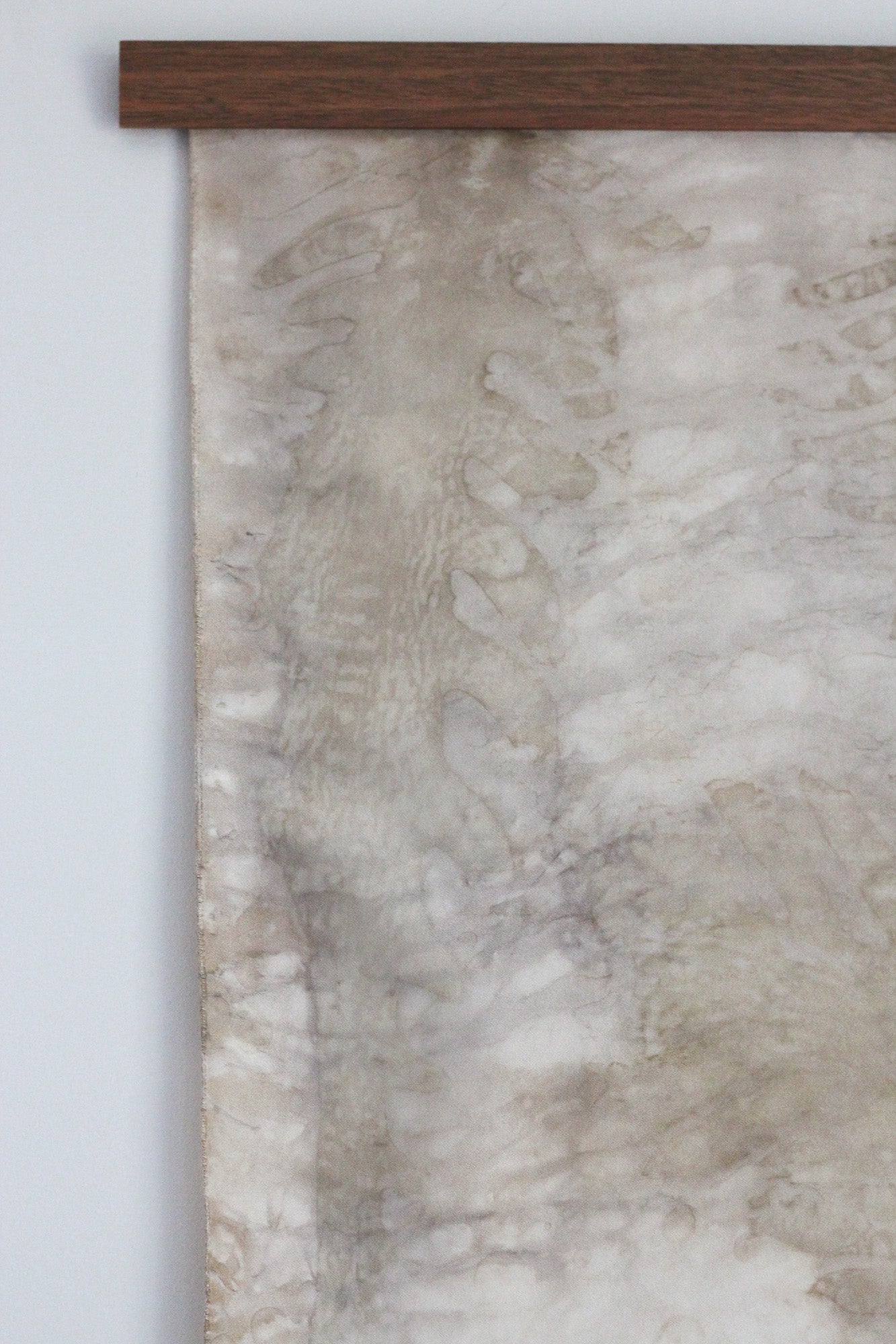 A close-up of a hanging textile wall hanging or scroll with a mottled beige, cream, and taupe pattern, suspended from a dark brown wooden rod against a light gray wall.