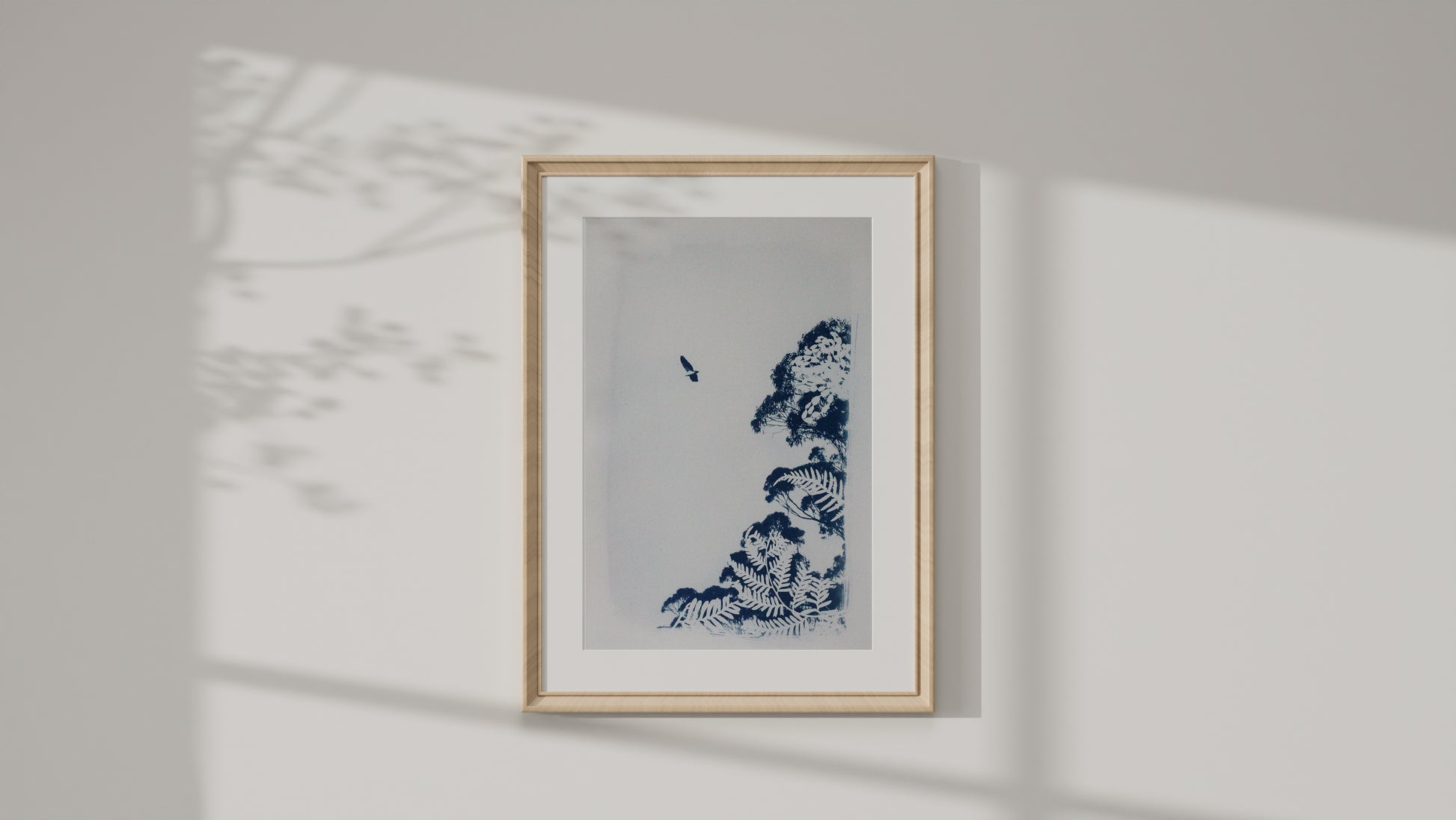 A framed art print featuring a cyanotype print of a sea eagle and tree on a white background, housed in a light wood frame and mounted on a white wall with soft shadow projections.