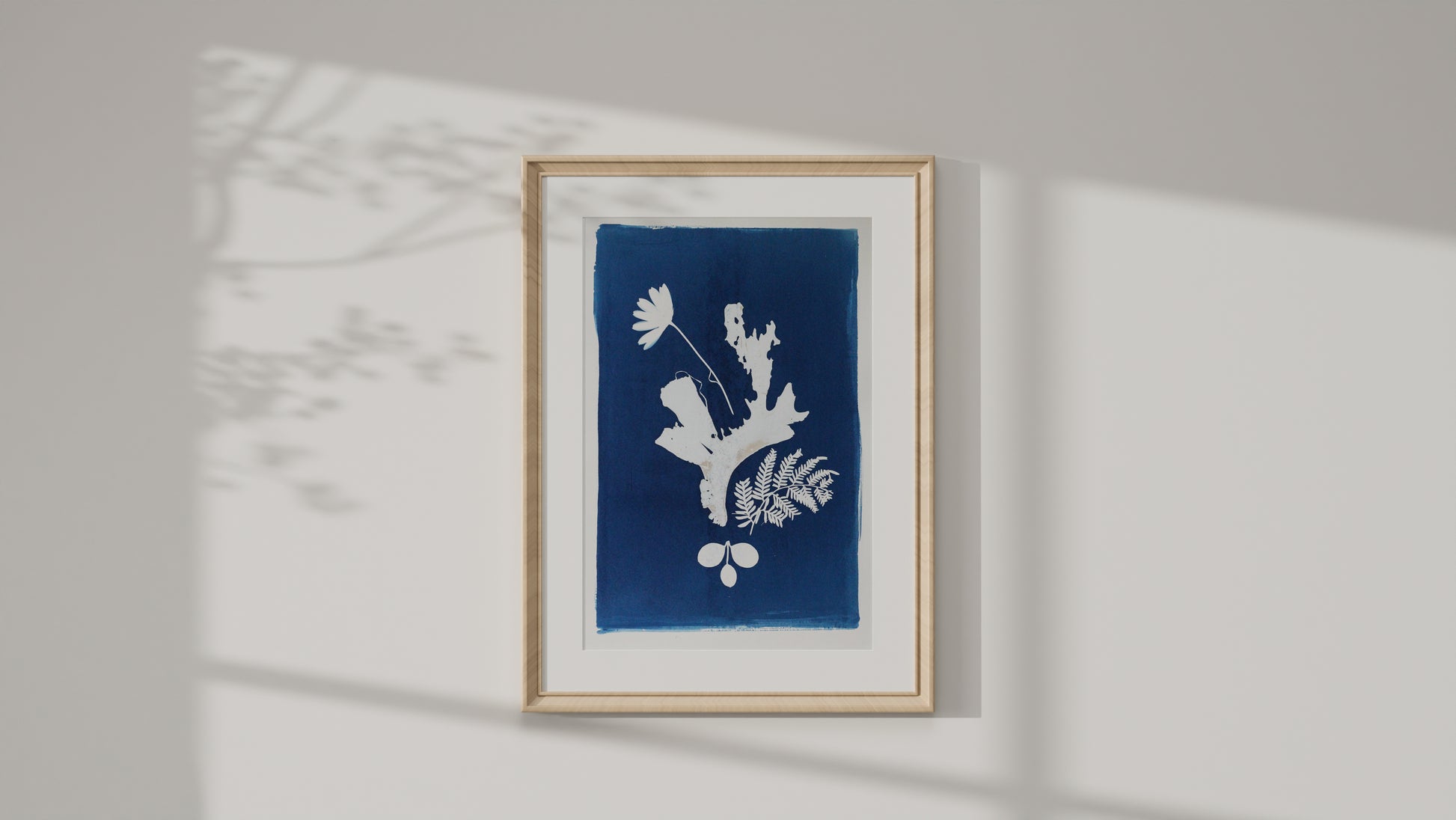 A framed botanical art cyanotype print with a blue background and white leafy shapes, mounted on a light gray wall with soft shadow projections.