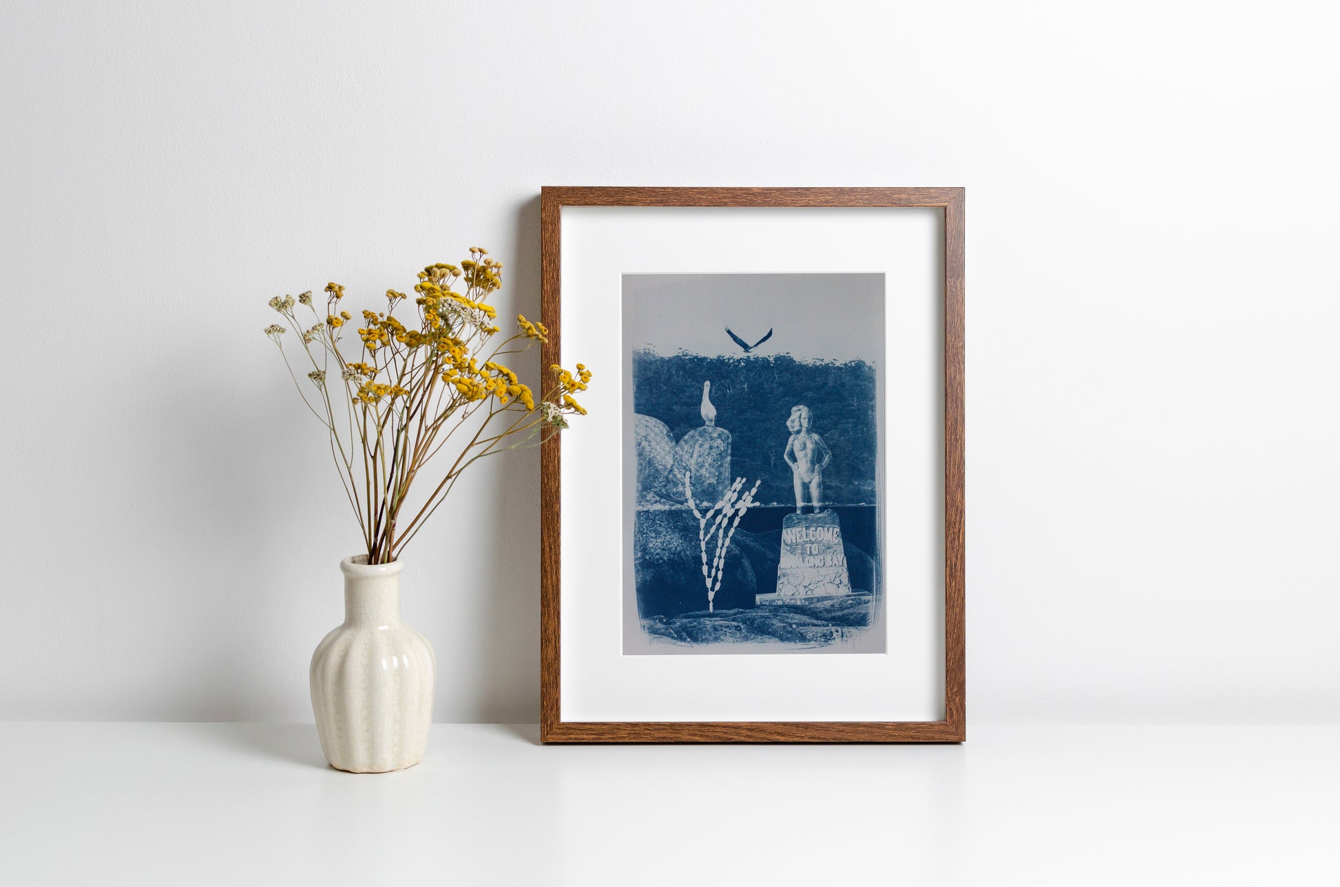 A framed art print with a blue and white cyanotype styled on a white surface next to a white ceramic vase with yellow flowers against a white wall.